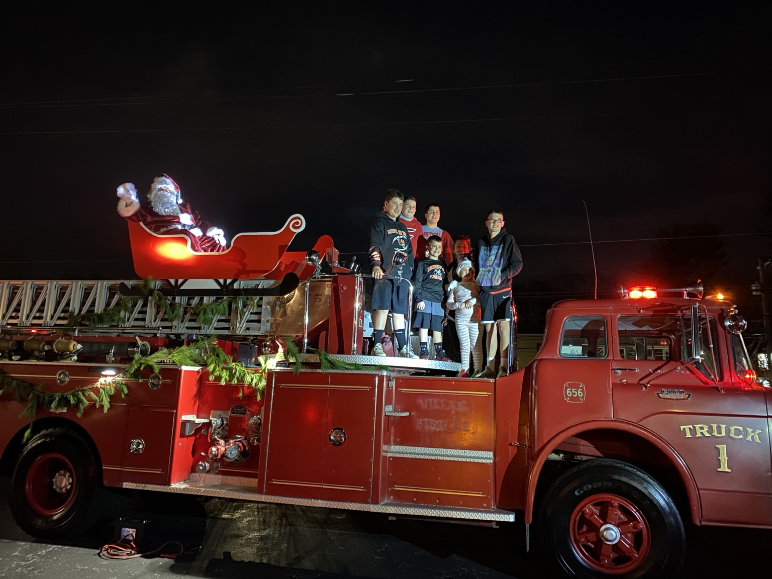 A restored red vintage Ford ladder fire truck at night, decorated with evergreen garland and a red sleigh mounted on the ladder rack, with a waving Santa in the sleigh and a group of riders standing on the rear step.